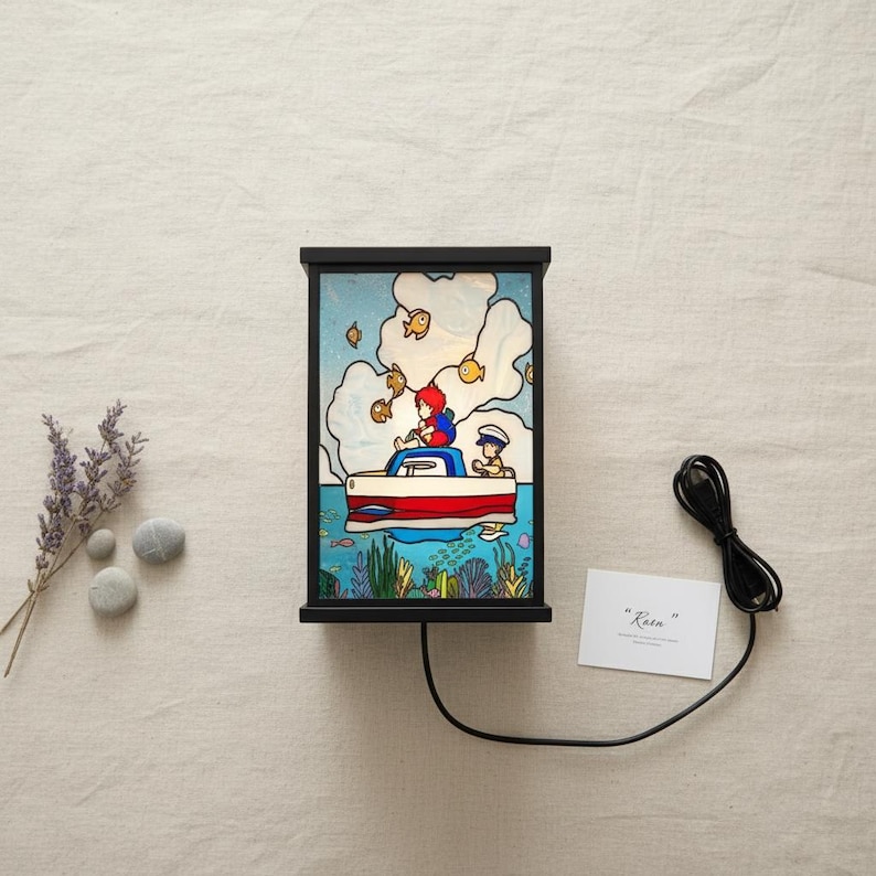 May include: A black framed lamp with a colorful illustration of a boat on the water with two people. The lamp has a red and white boat with a blue sky background and yellow fish. The text "Rain" is on a white card.