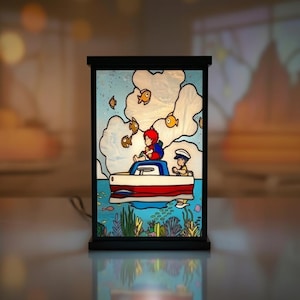 May include: A decorative stained glass lamp featuring a nautical scene. The lamp depicts two figures in a red and white boat, surrounded by fish and clouds. The base and frame are black, and the background is a light blue.