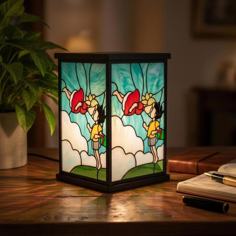 May include: A square stained glass lamp with a black frame. The lamp features a colorful design of children and flowers against a blue sky background. The lamp is sitting on a wooden table.