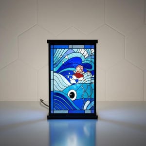May include: A stained glass lamp featuring a vibrant blue ocean scene with a cartoon character in a red outfit. The lamp has a black frame and base, and the image is illuminated from within.
