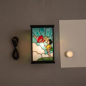 May include: A rectangular stained glass lamp with a black frame. The lamp features a colorful illustration of two children. One child is wearing a red dress and the other is wearing a yellow shirt. A black power cord is to the left of the lamp.