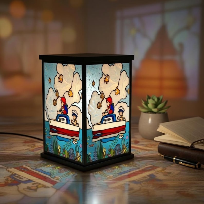 May include: A decorative lamp with a black frame features a colorful illustration of a boat with two people fishing. The lamp's design includes a blue sky, white clouds, and orange fish. The lamp is on a patterned surface.