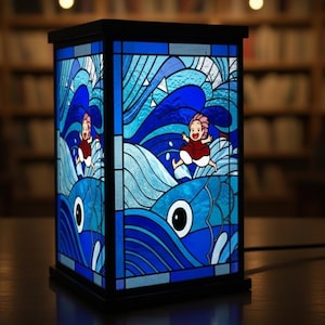 May include: A rectangular stained glass lamp with a black frame. The lamp features a blue ocean scene with a cartoon character in a red dress. The design includes waves and fish, with varying shades of blue and white.