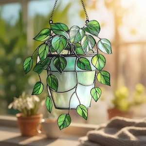 May include: A stained glass hanging planter featuring a trailing green plant. The planter is a light green colour with a geometric design, and the plant has vibrant green leaves. The planter hangs from a black chain, creating a decorative piece for a window or indoor space.