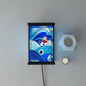 May include: A black-framed stained glass panel depicting a person in a red outfit riding a blue fish through blue waves. A white teacup and a lit candle sit nearby. The panel is plugged into an electrical outlet.