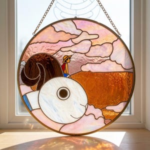 Könnte beinhalten: Ein rundes Buntglasfenster mit einer Cartoonfigur mit Strohhut und roter Weste, die auf einem braunen Objekt sitzt. Der Hintergrund zeigt rosa und orangefarbene Wolken und eine braune Landschaft. Das Fenster ist in Messing gerahmt und wird von einer Kette gehalten.