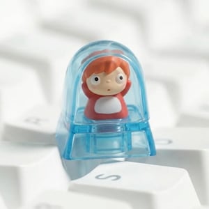 Op de afbeelding: Een aangepaste keycap met een stripfiguur in een transparante blauwe behuizing. Het personage heeft rood haar en een rode outfit met een witte slab. De keycap is ontworpen voor een computertoetsenbord.
