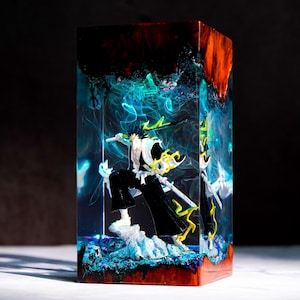 Op de afbeelding: Een rechthoekige hars sculptuur met een manga karakter in een dynamische pose. Het figuur is gekleed in zwart en wit, zwaaiend met een zwaard, tegen een achtergrond van blauwe en gele energie-effecten. Het harsblok heeft een rode en zwarte bovenkant.