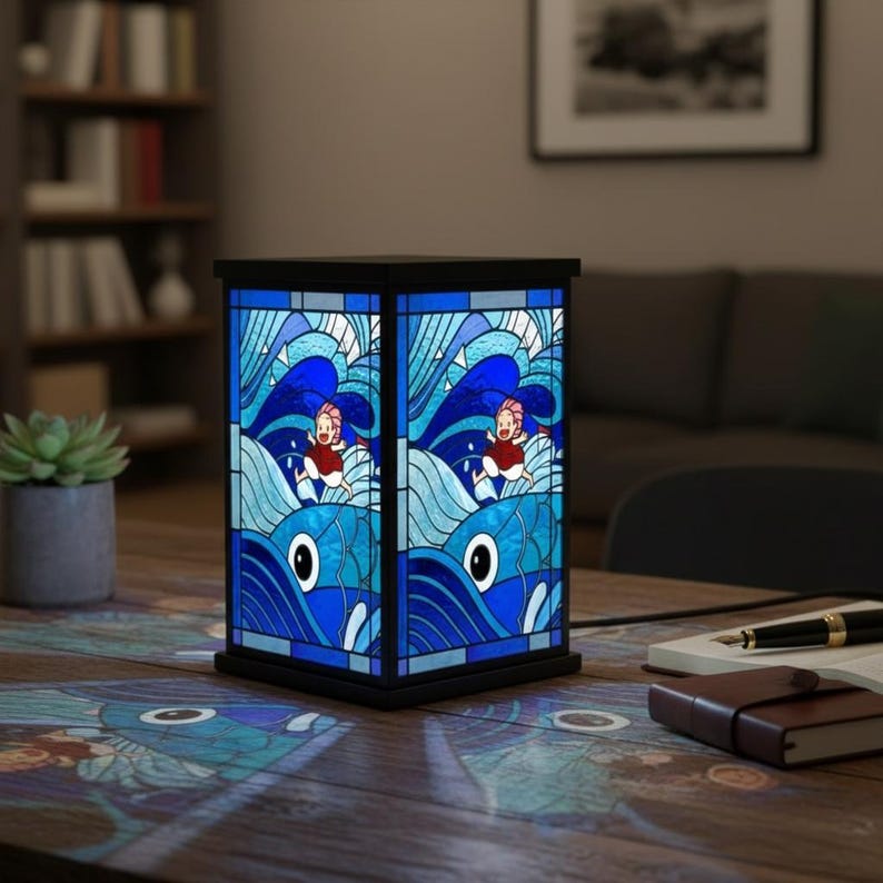 May include: A rectangular stained glass lamp with a black frame. The lamp features a blue and white ocean wave design with cartoon figures in red clothing. The lamp is on a wooden table with a notebook, pen, and a small plant.