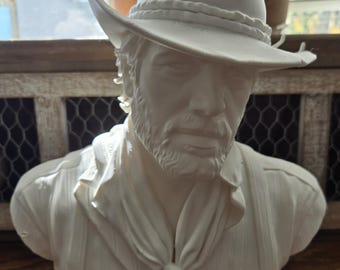 May include: A white matte bust sculpture of a bearded individual wearing a wide-brimmed hat, a neckerchief tied at the front, and a collared shirt with suspenders. The figure has a detailed facial expression with a short beard and mustache.