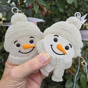 May include: Two white, glittery snowman ornaments featuring textured knit-style hats and scarves. Each snowman has black circular eyes, a curved black mouth, and a bright orange carrot-shaped nose. Silver metal loops are attached to the top.