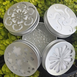 May include: Four silver-colored jar lids with decorative cut-out designs. The lids are arranged on a bed of green foliage. Each lid features a unique pattern, including floral and abstract shapes, adding a touch of elegance.
