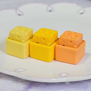 May include: Three colorful, square-shaped cheese-themed objects. Each cheese block sits atop a matching square container. The cheese blocks are yellow and orange, with holes. The containers are yellow and orange, arranged on a white plate.