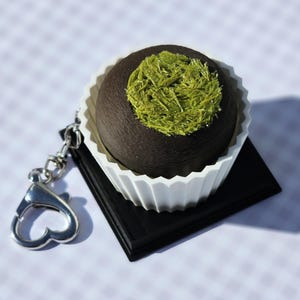May include: A dark brown sphere with a green textured topping, in a white fluted paper cup. It rests on a black square, with a silver keychain attached. The item is a decorative accessory.