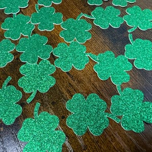 May include: A collection of green glitter shamrock cutouts scattered on a wooden surface. The four-leaf clovers are a vibrant green color and have a sparkling texture, perfect for St. Patrick's Day decorations.