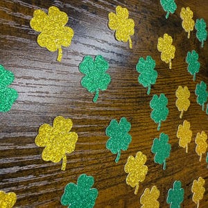 May include: Glittery green and gold shamrock cutouts scattered on a wooden surface. The four-leaf clovers are in various sizes and are ideal for St. Patrick's Day decorations or crafts. The glitter adds a festive touch.