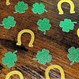 May include: A collection of St. Patrick's Day decorations, including green glitter shamrocks and gold glitter horseshoes, scattered on a dark wood-grain surface. The festive elements are arranged in a random pattern.