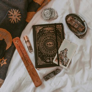 Könnte beinhalten: Eine Auswahl spiritueller Gegenstände auf einer weißen Oberfläche. Ein schwarzes Notizbuch mit goldenen Himmelsmustern, eine Tarotkarte, ein Räucherstäbchenhalter aus Holz, eine kleine Parfümflasche, ein Kristall, ein dekorativer Stein und eine Muschel mit Salbei.