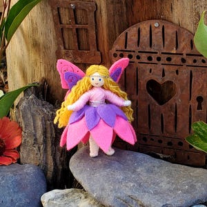 May include: A handmade felt fairy doll with pink and purple flower petals for a skirt, blonde yarn hair, and pink wings. The fairy is standing on a gray rock in front of a wooden door with a heart-shaped cutout.