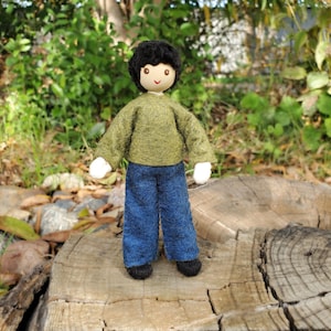 May include: A handmade doll with a wooden head, black hair, and a smiling face. The doll wears a green sweater, blue pants, white gloves, and black shoes. The doll is standing on a tree stump.