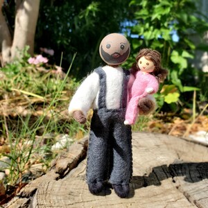 May include: A handmade felt doll of a man holding a smaller doll. The man doll has a painted face with a mustache and beard, wearing a white shirt, gray overalls, and black shoes. The smaller doll is wearing a pink outfit.