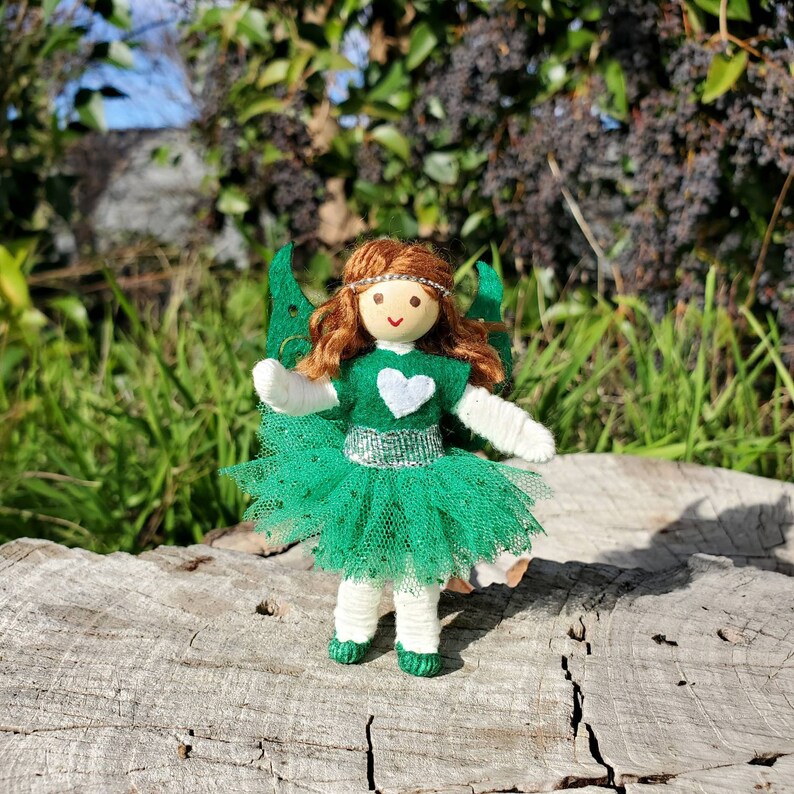 May include: A green fairy doll with a white heart on her dress and a green tulle skirt. She has brown hair and blue eyes. The doll is standing on a wooden surface.
