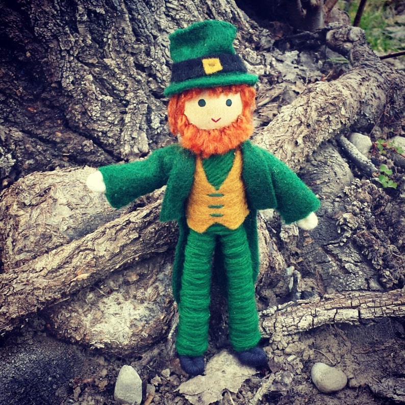A felt Irish doll dressed as a leprechaun with a green hat, orange beard, and green pants. The doll is standing in front of a tree trunk.
