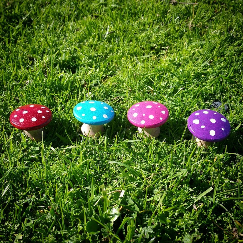 May include: Four painted wooden mushrooms with white polka dots. The mushrooms are painted in shades of pink, blue, purple, and red.