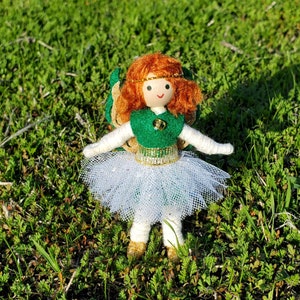 May include: A handmade felt doll with red hair, a green dress, and a white tulle skirt. The doll is wearing a gold crown and has a green felt flower on its chest. The doll is standing on a bed of green grass.