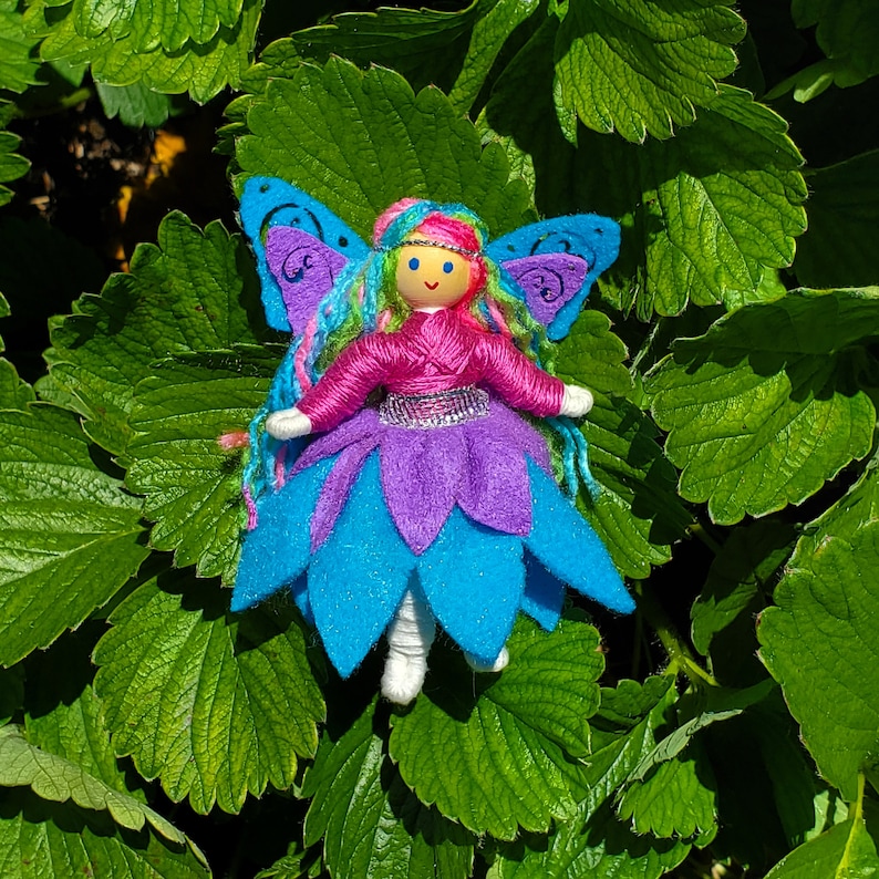 May include: A handmade felt fairy doll with blue, purple, and pink wings and a pink dress. The doll has a yarn hair and a painted face.