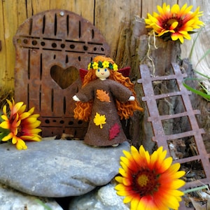 May include: A handmade felt doll with brown hair and a brown dress decorated with felt leaves. The doll is standing in front of a rusty metal gate and a ladder. There are also orange and yellow flowers in the image.