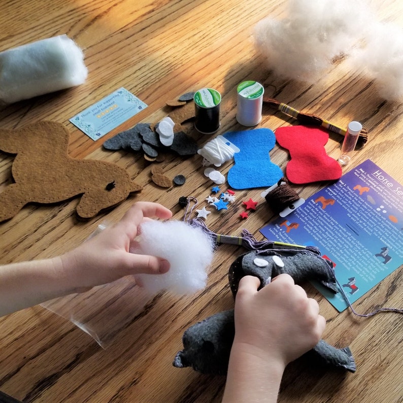 May include: A craft kit for making a felt horse. The kit includes felt pieces in brown, gray, blue, and red, along with stuffing, thread, and instructions. The kit also includes a small bag of white stars and a needle.