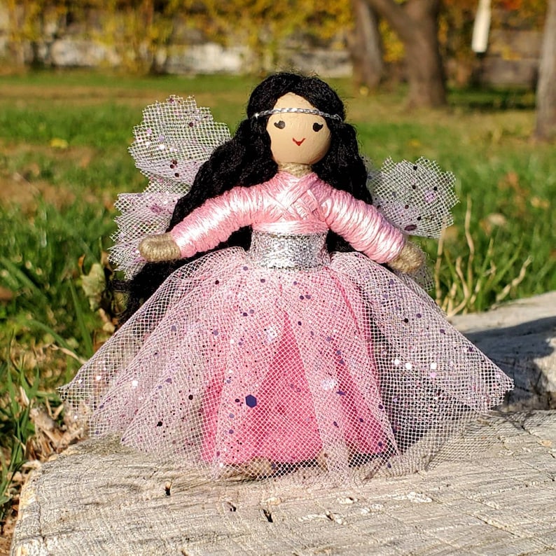 May include: A handmade fairy doll with long black hair, a pink dress with silver glitter, and a sheer pink tulle skirt. The doll has a painted face with brown eyes and a silver headband.