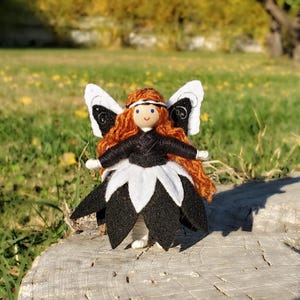 May include: A handmade felt fairy doll with black and white wings, a black dress, and a white flower-shaped skirt. The doll has red yarn hair and blue eyes.
