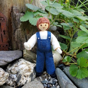 May include: A handmade doll with a wooden head and brown yarn hair. The doll wears a white shirt and blue denim overalls. The doll is standing on a bed of rocks with green foliage in the background.