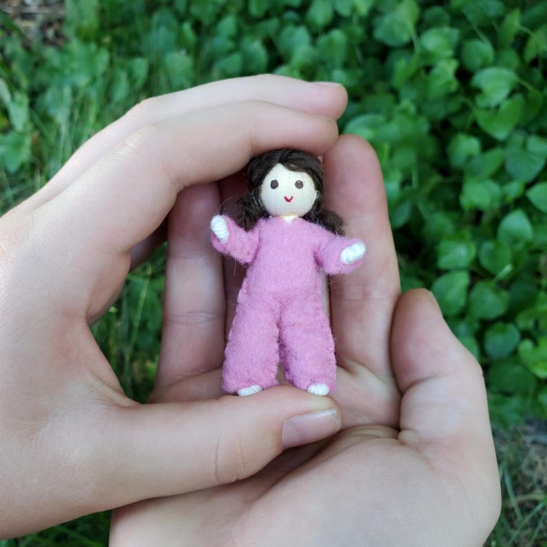 May include: A miniature doll with a pink jumpsuit and dark brown hair. The doll has a simple face with a small smile and white accents on the hands and feet. The doll is held in open hands against a background of green foliage.