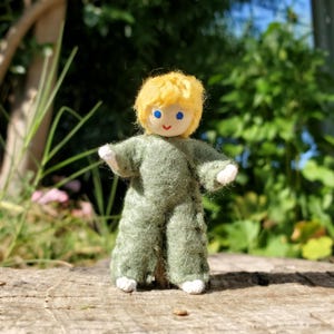 May include: A handmade doll with a yellow yarn hairstyle, blue eyes, and a red mouth. The doll wears a green jumpsuit with white accents on the hands and feet. The doll is standing on a wooden surface.
