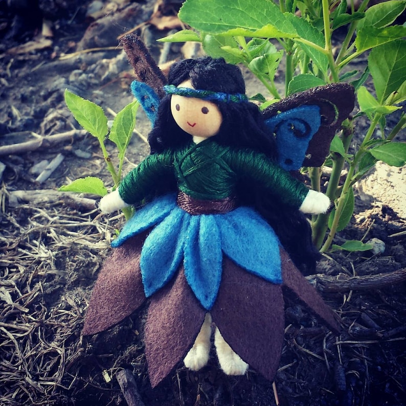 May include: A handmade felt fairy doll with brown, blue, and green fabric. The doll has long black hair, a green bodice, and a blue skirt with brown accents. The doll has a painted face with a blue headband.