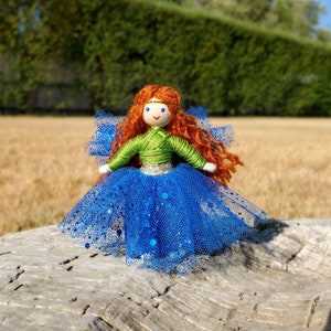 May include: A handmade doll with long, curly red hair, wearing a green shirt and a blue tulle skirt with glitter. The doll has a painted face with blue eyes and a smile.
