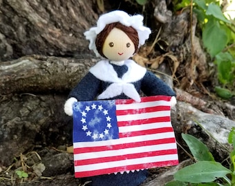 May include: A doll dressed in a blue and white sailor outfit holds a small American flag with 13 stars in a circle. The doll has brown hair and a white cap. The doll is standing in front of a tree trunk.