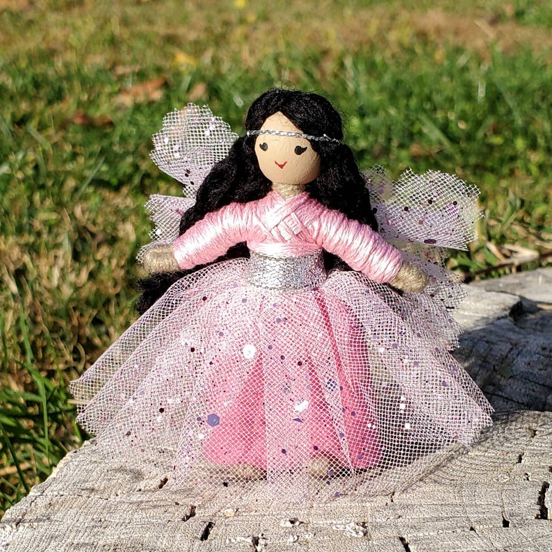 May include: A handmade doll with long black hair, wearing a pink tulle dress with silver glitter accents. The doll has a painted face with a headband and is holding a small bouquet of flowers.