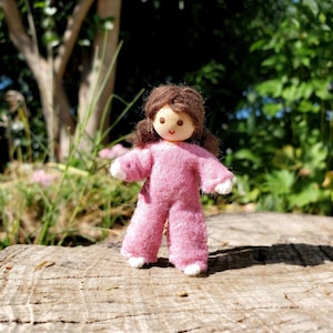May include: A handmade doll with a wooden head and brown hair, wearing a pink felt jumpsuit with white accents. The doll is standing on a wooden surface, with a blurred green background of foliage.