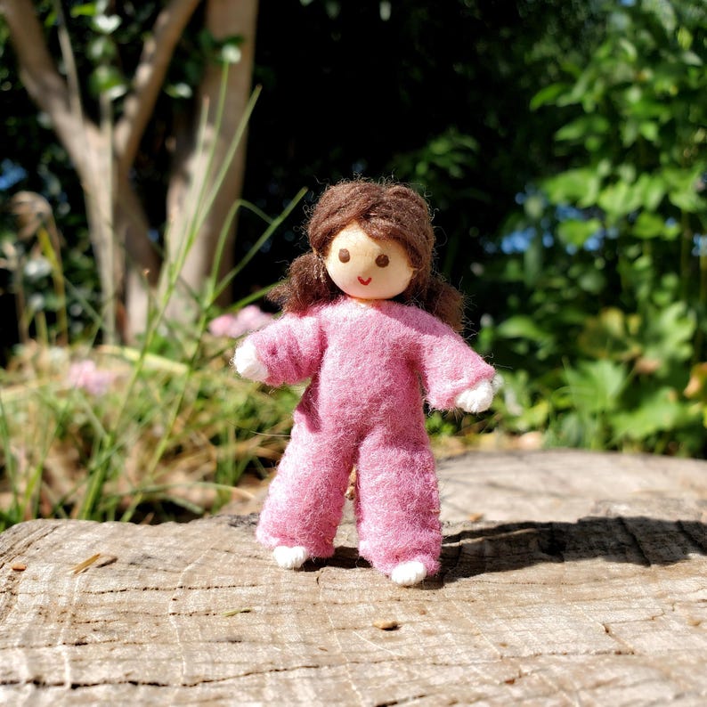 May include: A handmade doll with a wooden head and brown hair, wearing a pink felt jumpsuit with white accents. The doll is standing on a wooden surface, with a blurred green background of foliage.