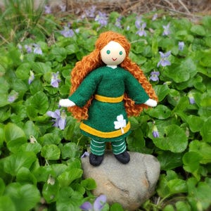May include: A handmade felt doll with curly red hair, wearing a green dress with a gold trim and a shamrock design. The doll is standing on a rock surrounded by green leaves and purple flowers.