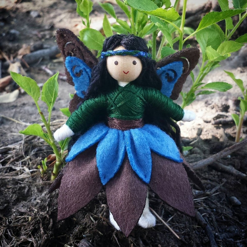 May include: A handmade felt fairy doll with brown hair, a green dress, blue wings, and a brown and blue flower skirt.