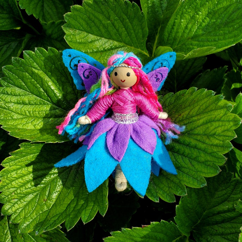 May include: A handmade felt fairy doll with pink yarn hair, blue wings, and a purple and blue skirt. The doll is sitting on a green leaf.