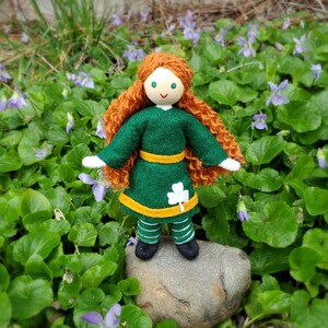May include: A handmade felt doll with red curly hair, a green dress with a shamrock, and a yellow belt. The doll is standing on a rock in a bed of green leaves and purple flowers.