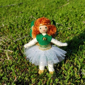 May include: A handmade doll with red yarn hair, a green felt dress, a white tulle skirt, and gold glitter accents. The doll is standing on a green grassy surface.