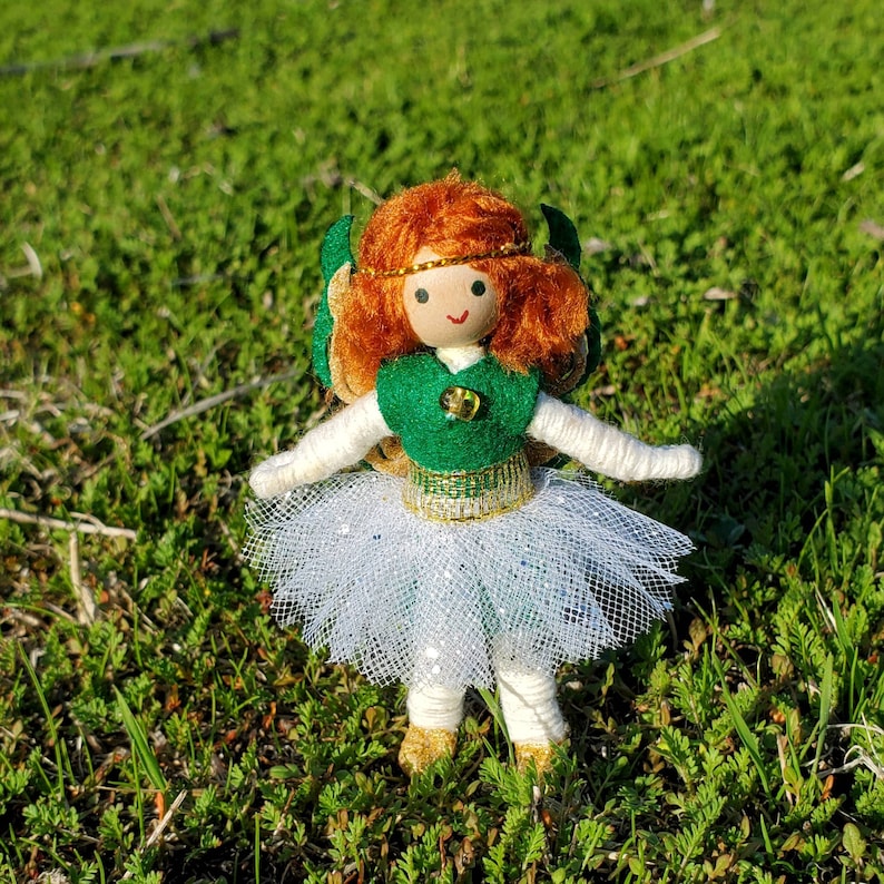 May include: A handmade doll with red yarn hair, a green felt dress, a white tulle skirt, and gold glitter accents. The doll is standing on a green grassy surface.