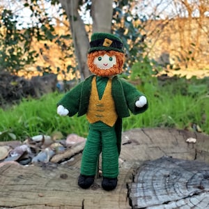 May include: A felt doll dressed as a leprechaun. The doll has a green jacket, a yellow vest, green pants, a green hat with a gold buckle, a red beard, and a friendly face.